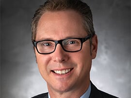 Daniel Ballin smiles at the camera in front of a gray background wearing black glasses.